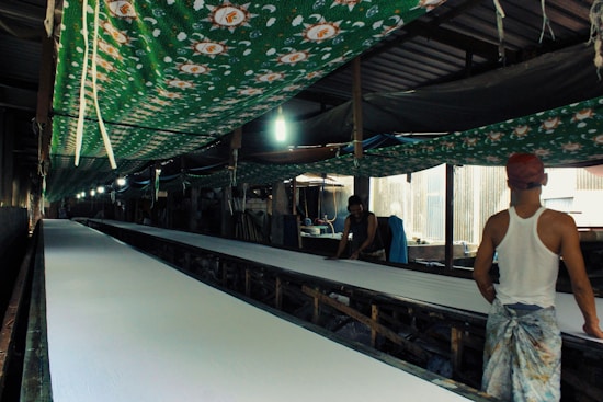 A long, narrow factory-like setting with two men working. They are handling large rolls of fabric on an extended table. The area is covered with green cloth decorated with patterns, hanging overhead. Strip lighting illuminates the space, creating shadows and highlights.