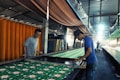 Two individuals are working together in a fabric printing workshop. They are engaged in hand-printing a long piece of fabric with a floral pattern. The workshop is lined with textiles, and a bright light shines in from the distance, illuminating part of the workspace. The environment appears industrial with a focus on traditional craftsmanship.