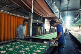 Two individuals are working together in a fabric printing workshop. They are engaged in hand-printing a long piece of fabric with a floral pattern. The workshop is lined with textiles, and a bright light shines in from the distance, illuminating part of the workspace. The environment appears industrial with a focus on traditional craftsmanship.
