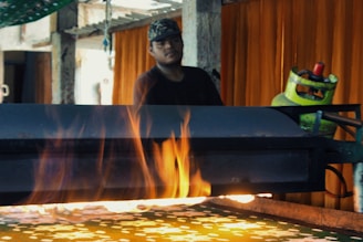 A person is standing behind a table on which a machine emits visible flames. The setting appears to be industrial, with metal and mechanical equipment, including a propane gas cylinder. The background features orange curtains and some hanging items, suggesting an indoor environment.