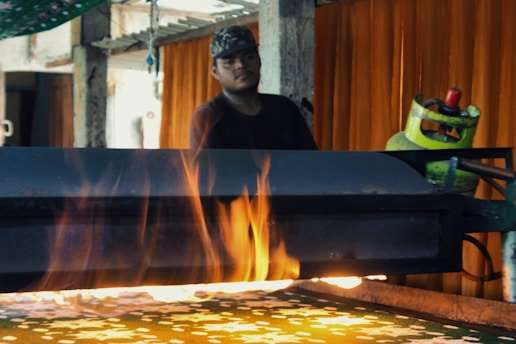 A person is standing behind a table on which a machine emits visible flames. The setting appears to be industrial, with metal and mechanical equipment, including a propane gas cylinder. The background features orange curtains and some hanging items, suggesting an indoor environment.