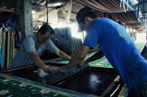 Two men are engaged in a manual screen-printing process in an industrial setting. They are using large screens to apply patterns onto fabrics. The environment appears to be a workshop with various tools and materials scattered around. Ambient lighting creates a focused but slightly dim atmosphere.