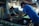 Two men are engaged in a manual screen-printing process in an industrial setting. They are using large screens to apply patterns onto fabrics. The environment appears to be a workshop with various tools and materials scattered around. Ambient lighting creates a focused but slightly dim atmosphere.
