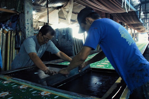 Two men are engaged in a manual screen-printing process in an industrial setting. They are using large screens to apply patterns onto fabrics. The environment appears to be a workshop with various tools and materials scattered around. Ambient lighting creates a focused but slightly dim atmosphere.