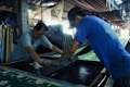 Two men are engaged in a manual screen-printing process in an industrial setting. They are using large screens to apply patterns onto fabrics. The environment appears to be a workshop with various tools and materials scattered around. Ambient lighting creates a focused but slightly dim atmosphere.