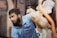 A man with a beard in a blue polo shirt holding a live chicken with one hand. The background appears to be an outdoor setting, slightly blurred, with various colors and shapes indicating other objects or structures.
