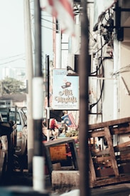A street scene featuring a restaurant sign reading 'Ali Mustafa' with various objects like a wooden pallet and a box in the foreground. The background includes a Malaysian flag, an air conditioning unit, and a parked vehicle.