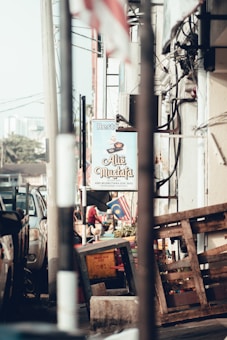A street scene featuring a restaurant sign reading 'Ali Mustafa' with various objects like a wooden pallet and a box in the foreground. The background includes a Malaysian flag, an air conditioning unit, and a parked vehicle.