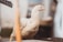 A white chicken standing on a wooden surface, with a slightly blurry background. The chicken is looking slightly to the side, and there is an out-of-focus object in the foreground, possibly a piece of wood or metal.