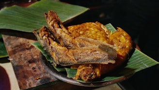 A plate of deep-fried fish and fried food items is arranged on banana leaves. The crispy texture and golden brown color suggest they are freshly cooked. The arrangement is on a wooden surface, indicating a rustic presentation.