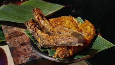 A plate of deep-fried fish and fried food items is arranged on banana leaves. The crispy texture and golden brown color suggest they are freshly cooked. The arrangement is on a wooden surface, indicating a rustic presentation.