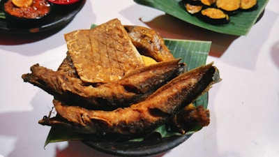 A dish featuring crispy fried fish and a rectangular piece of tempeh placed on a green banana leaf. The background shows small plates with additional food items, including sliced vegetables.