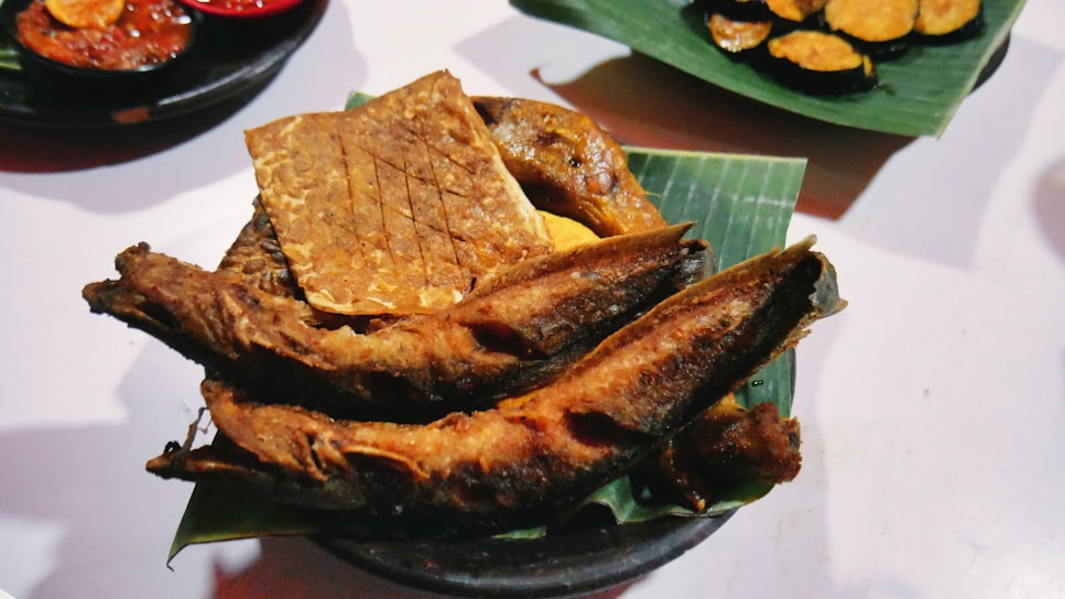 A dish featuring crispy fried fish and a rectangular piece of tempeh placed on a green banana leaf. The background shows small plates with additional food items, including sliced vegetables.