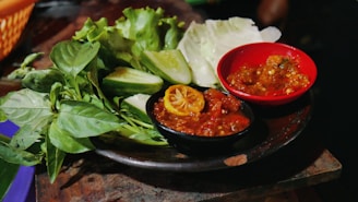A plate arranged with fresh green vegetables, including leafy greens and cucumber slices. Two small bowls of vibrant red chili sauce are placed on the plate, adding a pop of color and spice. The atmosphere suggests a rustic, flavorful, and possibly homemade meal presentation.