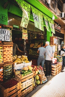 A bustling fruit and vegetable market with crates of bananas, limes, oranges, and various other produce stacked at the entrance. Two people are browsing the selection, while colorful signs with prices hang above them. The storefront is vibrant with a green exterior.