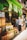 A bustling fruit and vegetable market with crates of bananas, limes, oranges, and various other produce stacked at the entrance. Two people are browsing the selection, while colorful signs with prices hang above them. The storefront is vibrant with a green exterior.