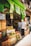 A bustling fruit and vegetable market with crates of bananas, limes, oranges, and various other produce stacked at the entrance. Two people are browsing the selection, while colorful signs with prices hang above them. The storefront is vibrant with a green exterior.