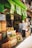 A bustling fruit and vegetable market with crates of bananas, limes, oranges, and various other produce stacked at the entrance. Two people are browsing the selection, while colorful signs with prices hang above them. The storefront is vibrant with a green exterior.