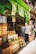 A bustling fruit and vegetable market with crates of bananas, limes, oranges, and various other produce stacked at the entrance. Two people are browsing the selection, while colorful signs with prices hang above them. The storefront is vibrant with a green exterior.