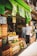 A bustling fruit and vegetable market with crates of bananas, limes, oranges, and various other produce stacked at the entrance. Two people are browsing the selection, while colorful signs with prices hang above them. The storefront is vibrant with a green exterior.
