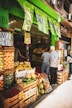 A bustling fruit and vegetable market with crates of bananas, limes, oranges, and various other produce stacked at the entrance. Two people are browsing the selection, while colorful signs with prices hang above them. The storefront is vibrant with a green exterior.