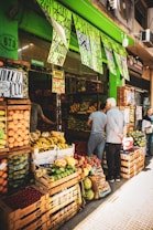 A bustling fruit and vegetable market with crates of bananas, limes, oranges, and various other produce stacked at the entrance. Two people are browsing the selection, while colorful signs with prices hang above them. The storefront is vibrant with a green exterior.