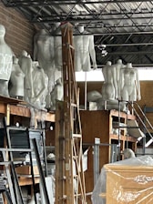 A cluttered storage space containing numerous white mannequins arranged on shelves. A wooden ladder is positioned in the center amidst other items such as boards, paper, and packaging material. The room has an industrial appearance with metal ceiling beams and a brick wall.