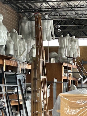 A cluttered storage space containing numerous white mannequins arranged on shelves. A wooden ladder is positioned in the center amidst other items such as boards, paper, and packaging material. The room has an industrial appearance with metal ceiling beams and a brick wall.