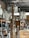 A cluttered storage space containing numerous white mannequins arranged on shelves. A wooden ladder is positioned in the center amidst other items such as boards, paper, and packaging material. The room has an industrial appearance with metal ceiling beams and a brick wall.