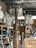 A cluttered storage space containing numerous white mannequins arranged on shelves. A wooden ladder is positioned in the center amidst other items such as boards, paper, and packaging material. The room has an industrial appearance with metal ceiling beams and a brick wall.