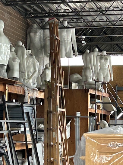 A cluttered storage space containing numerous white mannequins arranged on shelves. A wooden ladder is positioned in the center amidst other items such as boards, paper, and packaging material. The room has an industrial appearance with metal ceiling beams and a brick wall.