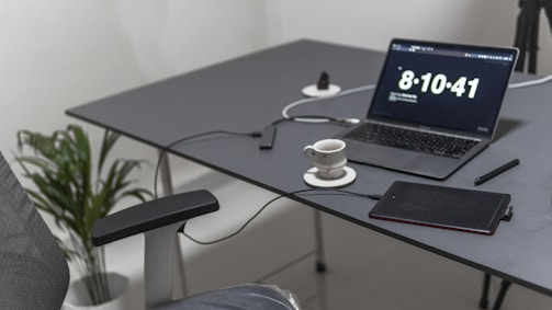 A minimalist workspace features a gray desk with a laptop showing the time, a cup of coffee on a saucer, a stylus pen, and a black electronic device connected by a cable. A plant in a white pot is placed beside the desk, and an office chair with a fabric backrest is partially visible.