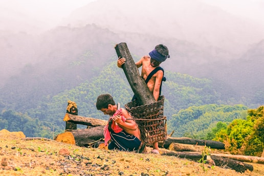 Two individuals are engaged in manual labor, carrying large logs in a mountainous terrain. One person is bent over under the weight of a log, while the other stands near with a log resting inside a woven basket strapped to their back. The background features misty mountains and lush greenery, creating a dramatic natural setting.