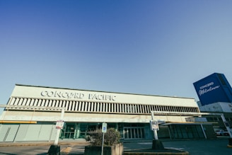 A large building with 'Concord Pacific' written prominently on its facade. The architecture features vertical slats above and below the signage. On the right side, a tall blue structure with 'Concord Metrotown' is visible. The area appears to be a parking lot with signs posted near the entrance.