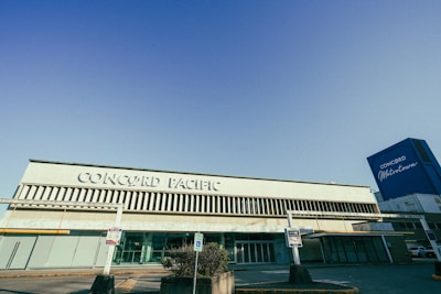 A large building with 'Concord Pacific' written prominently on its facade. The architecture features vertical slats above and below the signage. On the right side, a tall blue structure with 'Concord Metrotown' is visible. The area appears to be a parking lot with signs posted near the entrance.
