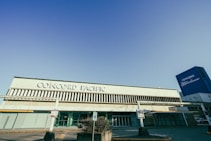 A large building with 'Concord Pacific' written prominently on its facade. The architecture features vertical slats above and below the signage. On the right side, a tall blue structure with 'Concord Metrotown' is visible. The area appears to be a parking lot with signs posted near the entrance.