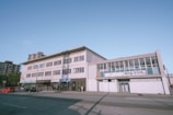A street view of a multi-story commercial building displaying signs in both English and Korean. The building features large windows and is situated along a relatively quiet road with a few parked cars and a couple of people walking nearby. In the background, there are a few taller residential buildings and a clear blue sky.