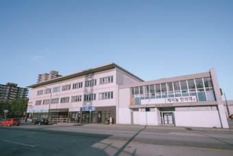 A street view of a multi-story commercial building displaying signs in both English and Korean. The building features large windows and is situated along a relatively quiet road with a few parked cars and a couple of people walking nearby. In the background, there are a few taller residential buildings and a clear blue sky.