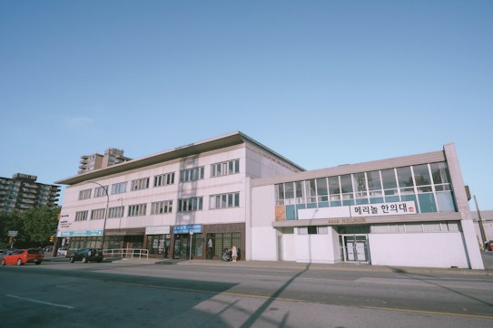 A street view of a multi-story commercial building displaying signs in both English and Korean. The building features large windows and is situated along a relatively quiet road with a few parked cars and a couple of people walking nearby. In the background, there are a few taller residential buildings and a clear blue sky.