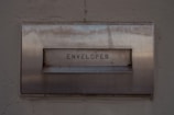 A metal mailbox with the word 'ENVELOPES' engraved on its front panel, set against a textured concrete wall. The surface shows signs of age with some rust marks.