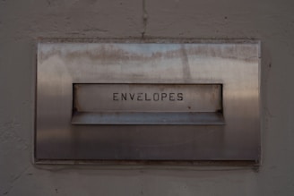 A metal mailbox with the word 'ENVELOPES' engraved on its front panel, set against a textured concrete wall. The surface shows signs of age with some rust marks.