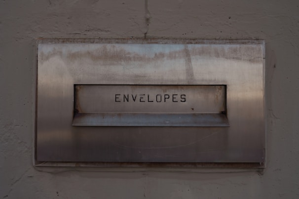 A metal mailbox with the word 'ENVELOPES' engraved on its front panel, set against a textured concrete wall. The surface shows signs of age with some rust marks.