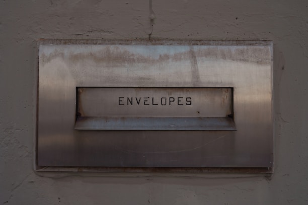 A metal mailbox with the word 'ENVELOPES' engraved on its front panel, set against a textured concrete wall. The surface shows signs of age with some rust marks.
