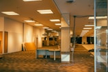 An empty, large indoor space with exposed ceiling lights and a patterned floor. There are various pieces of furniture and materials strewn around, including a desk with wires extending from it. A sign indicating a fitting room is visible in the background. The area appears to be in the process of being dismantled or set up.