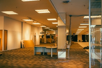 An empty, large indoor space with exposed ceiling lights and a patterned floor. There are various pieces of furniture and materials strewn around, including a desk with wires extending from it. A sign indicating a fitting room is visible in the background. The area appears to be in the process of being dismantled or set up.