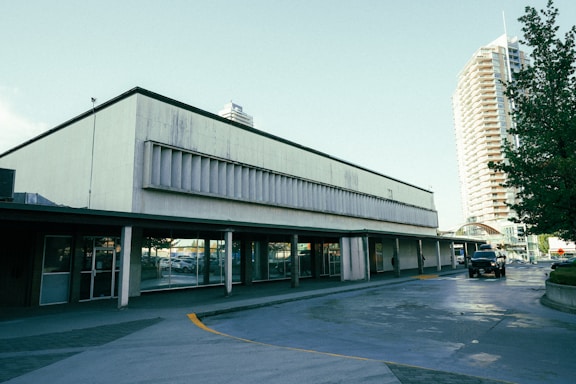 A commercial building with large glass windows and a concrete facade is situated next to a street. The structure appears somewhat aged with signs of wear on the exterior. In the background, a tall residential or office building with numerous windows is visible. Several cars, including a black vehicle, are parked nearby, and large green trees are present, adding some foliage to the urban setting.