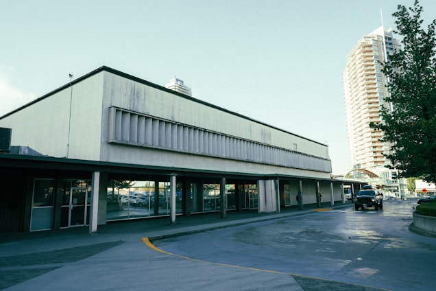 A commercial building with large glass windows and a concrete facade is situated next to a street. The structure appears somewhat aged with signs of wear on the exterior. In the background, a tall residential or office building with numerous windows is visible. Several cars, including a black vehicle, are parked nearby, and large green trees are present, adding some foliage to the urban setting.