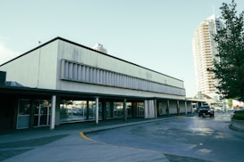 A commercial building with large glass windows and a concrete facade is situated next to a street. The structure appears somewhat aged with signs of wear on the exterior. In the background, a tall residential or office building with numerous windows is visible. Several cars, including a black vehicle, are parked nearby, and large green trees are present, adding some foliage to the urban setting.