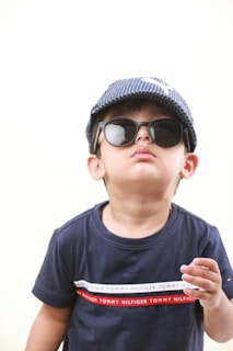 A young child wearing dark sunglasses and a blue-striped cap stands against a light background. The child is also wearing a dark blue t-shirt with a red and white stripe across the chest that contains the text 'Tommy Hilfiger'. The expression appears relaxed and confident.