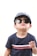 A young child wearing dark sunglasses and a blue-striped cap stands against a light background. The child is also wearing a dark blue t-shirt with a red and white stripe across the chest that contains the text 'Tommy Hilfiger'. The expression appears relaxed and confident.
